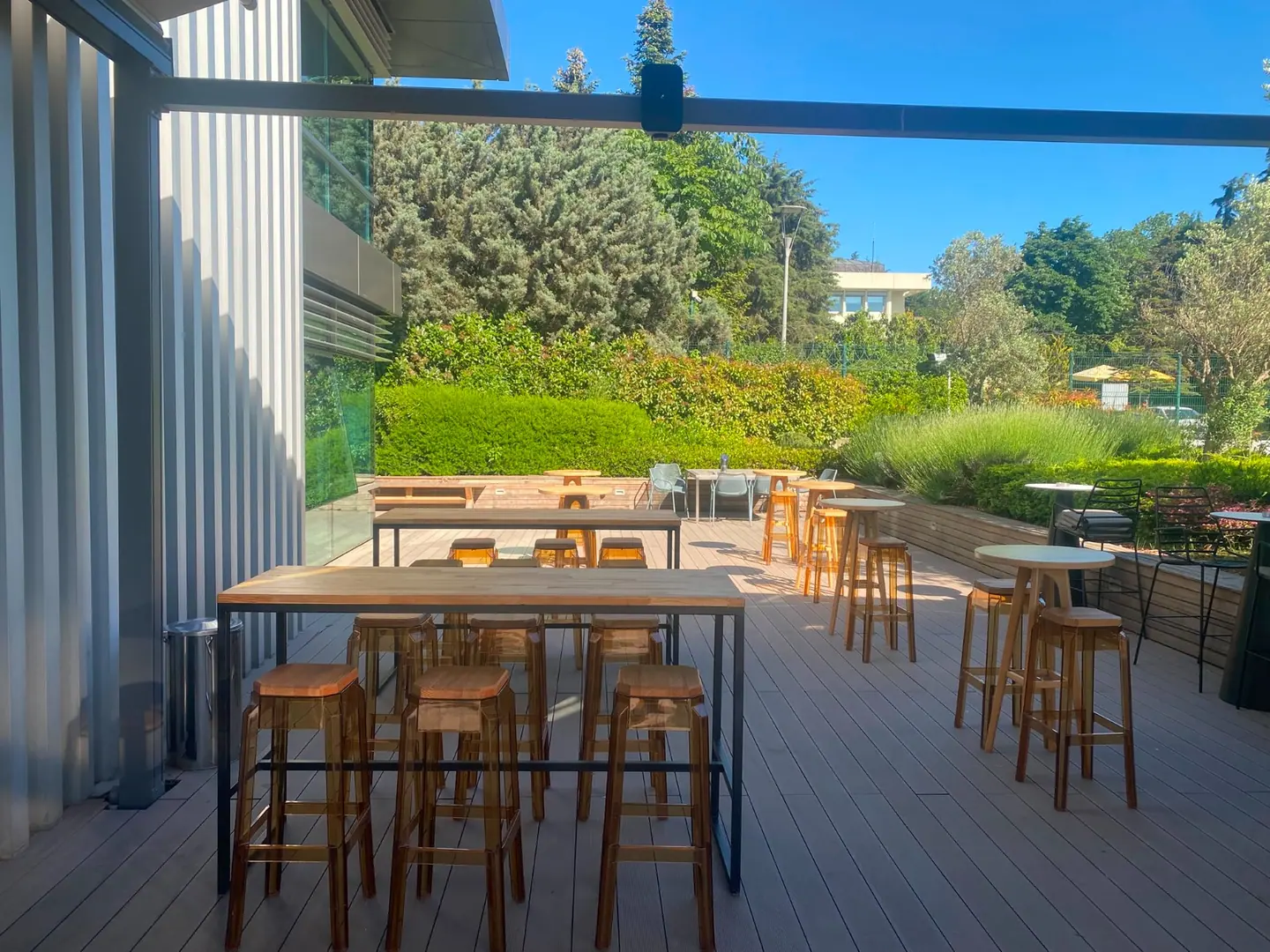 Ağaçlı açık terasta ahşap bistro masa ve taburelerin geniş açılı yerleşimi